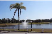 Pond with a central water fountain, a palm tree, lamp posts, and a white fence under a clear blue sky.