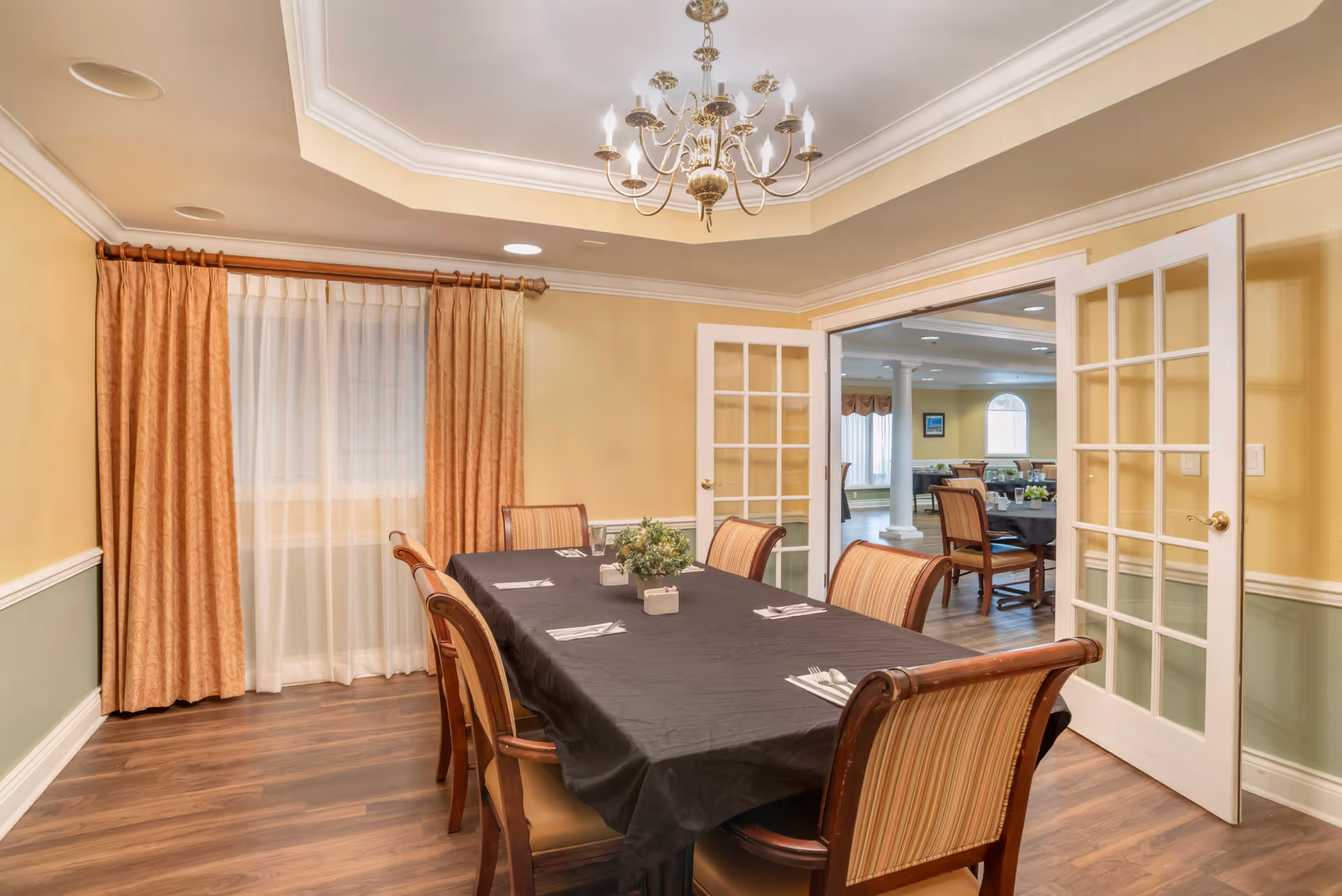 A dining room with a rectangular table covered with a black tablecloth, set with napkins and silverware. Six wooden chairs with striped upholstery surround the table. The room has yellow walls with white trim, a window with sheer white curtains and orange drapes, and a chandelier hanging from a tray ceiling. French doors open to another room with more tables and chairs.