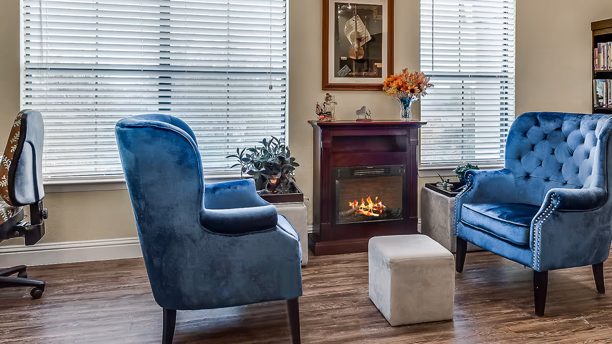 Cozy seating area with two blue armchairs, a small ottoman, and an electric fireplace between two large windows.