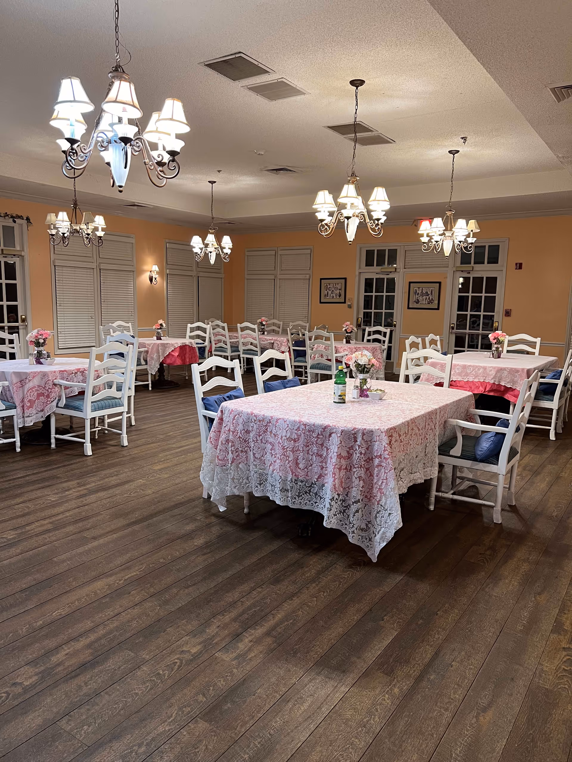 A dining room with multiple tables covered in pink and white lace tablecloths. Each table is surrounded by white wooden chairs with blue cushions. Small flower arrangements are placed on each table. The room has warm yellow walls, wooden flooring, and several chandeliers hanging from the ceiling providing soft lighting. Windows with closed blinds and framed pictures are visible on the walls.