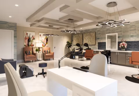 A modern and spacious salon area in a senior living facility featuring multiple styling stations with chairs and mirrors, a white manicure table with chairs in the foreground, two comfortable armchairs with floral artwork on the wall, and decorative lighting fixtures hanging from a coffered ceiling.