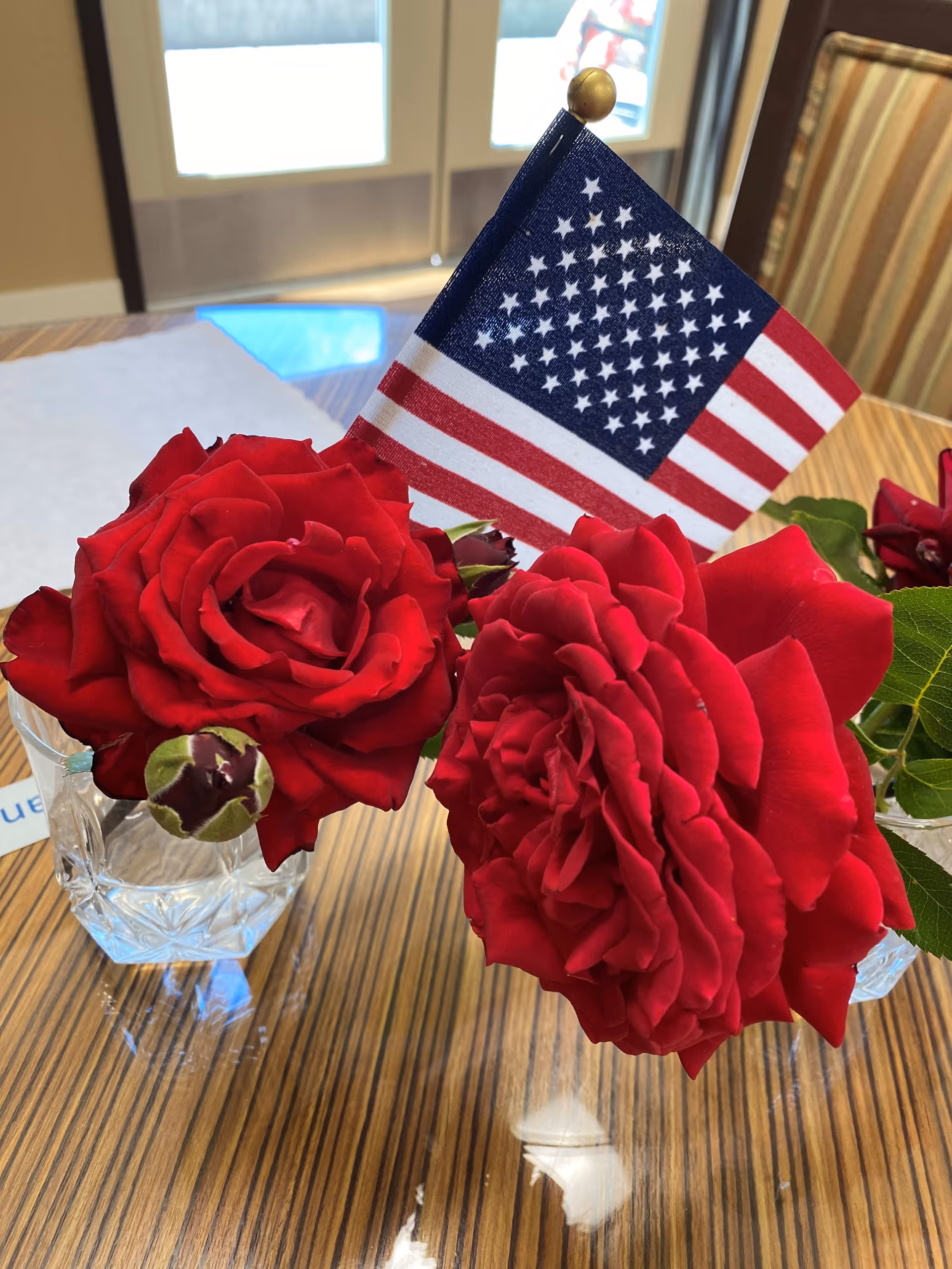 Close-up of two red roses in small glass vases on a wooden table with an American flag placed between them. In the background, there is a window and part of a striped chair.