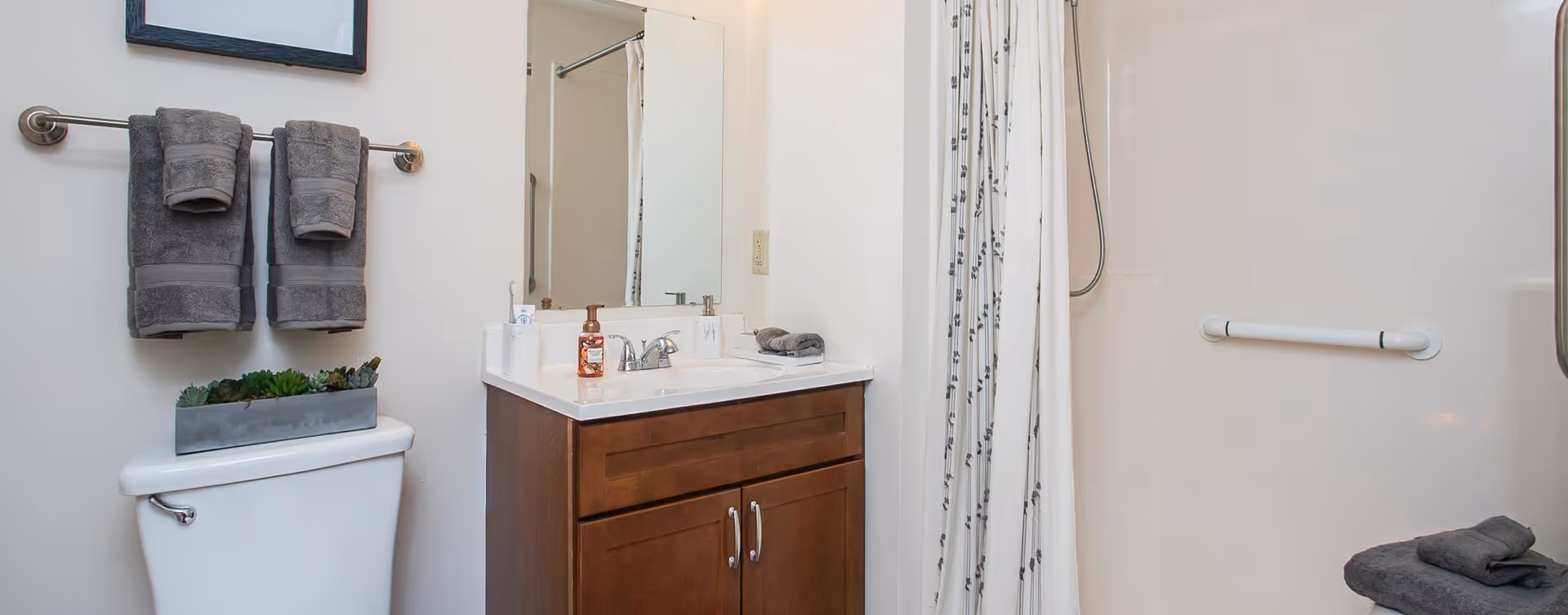 A well-lit bathroom featuring a wooden vanity with a sink, a large mirror, and neatly arranged towels. The shower area is visible with a curtain, and a small planter with succulents is placed on the vanity.
