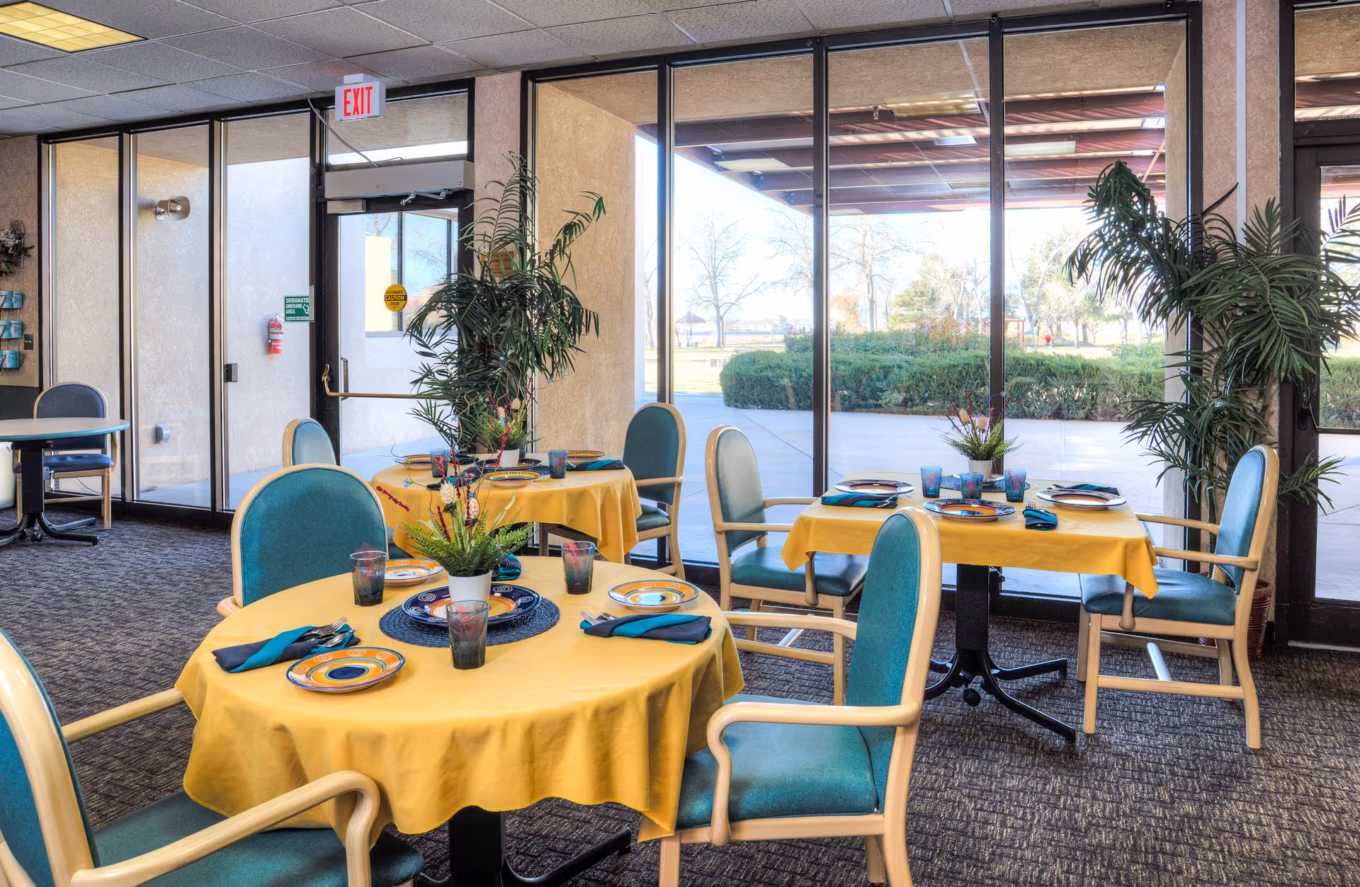 Dining area with round tables covered in yellow tablecloths, each set with plates, glasses, and teal napkins. The chairs have teal cushions and light wooden frames. Large windows and glass doors provide a view of an outdoor patio and greenery. Potted plants are placed near the windows and on the tables.