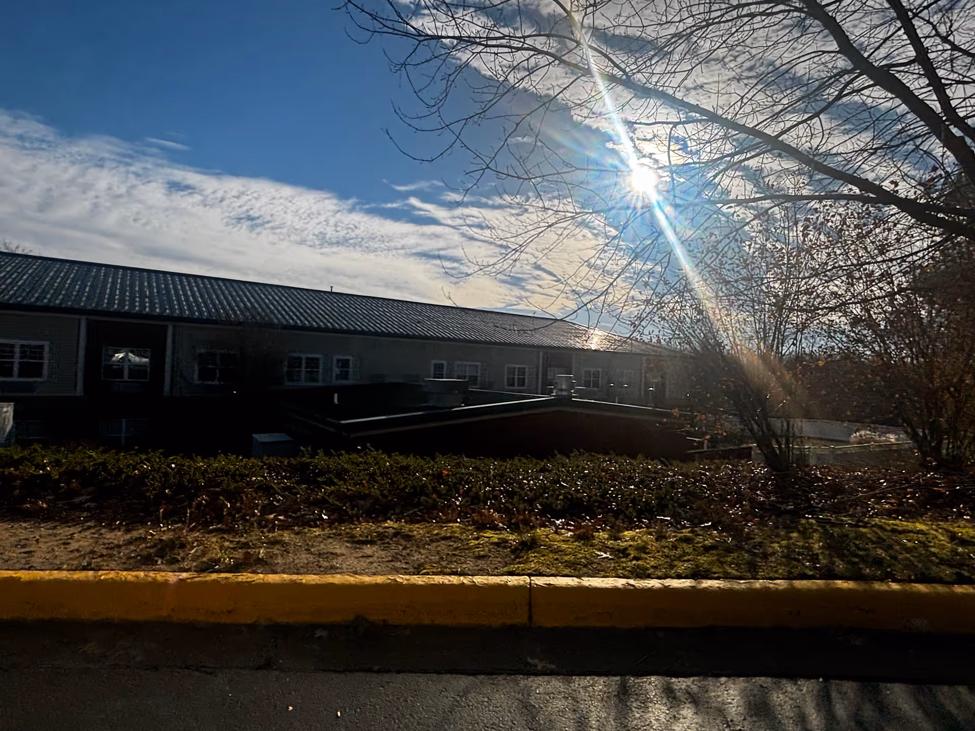 Exterior view of a senior living facility building with a metal roof under a partly cloudy sky. The sun is shining brightly through bare tree branches, casting light and shadows on the ground and bushes in front of the building.