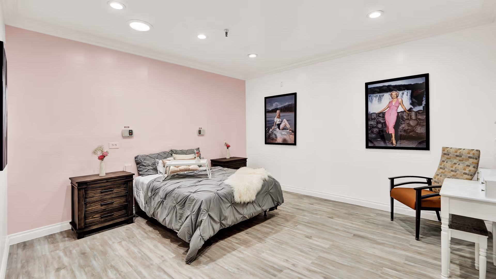 A bedroom with a bed covered in gray bedding and a white fur throw. There are two dark wooden nightstands with small flower vases on each side of the bed. The walls are painted white and light pink, with two framed pictures of women in vintage-style dresses hanging on the white wall. A patterned armchair and a white desk with a small stool are positioned on the right side of the room. The floor is light wood, and recessed ceiling lights illuminate the space.