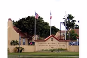 Exterior view of Spanish Meadows Senior Meadows Assisted Living facility with a beige wall and sign displaying the facility name, three flagpoles with American, Texas, and another flag, palm trees, and a building with a chimney in the background.