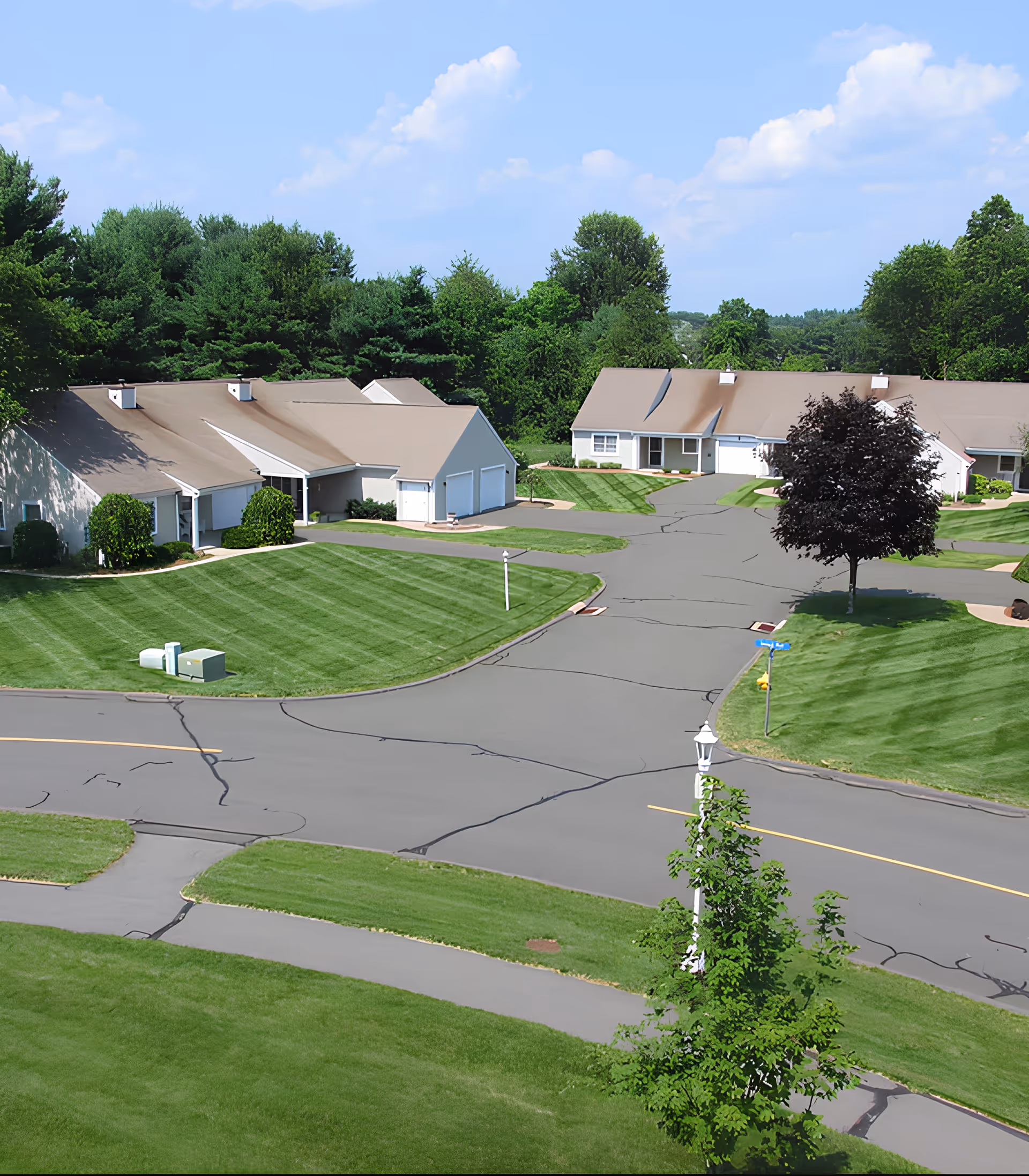 Aerial view of a residential community with single-story houses featuring beige roofs, well-maintained green lawns, paved roads, and surrounded by trees under a blue sky with scattered clouds.