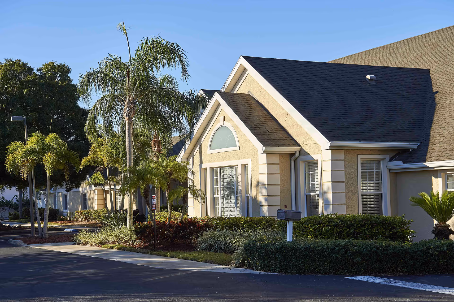 Exterior view of a residential building with beige walls, white trim, and a dark shingled roof. The building is surrounded by well-maintained landscaping including palm trees, bushes, and other greenery. The scene is lit by sunlight under a clear blue sky.