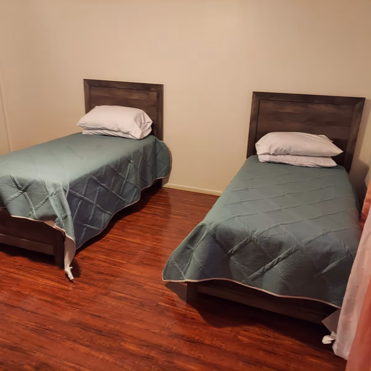 A simple bedroom with two single beds placed parallel to each other. Each bed has a wooden headboard, a white pillow, and a green quilted bedspread. The room has wooden flooring and plain beige walls.