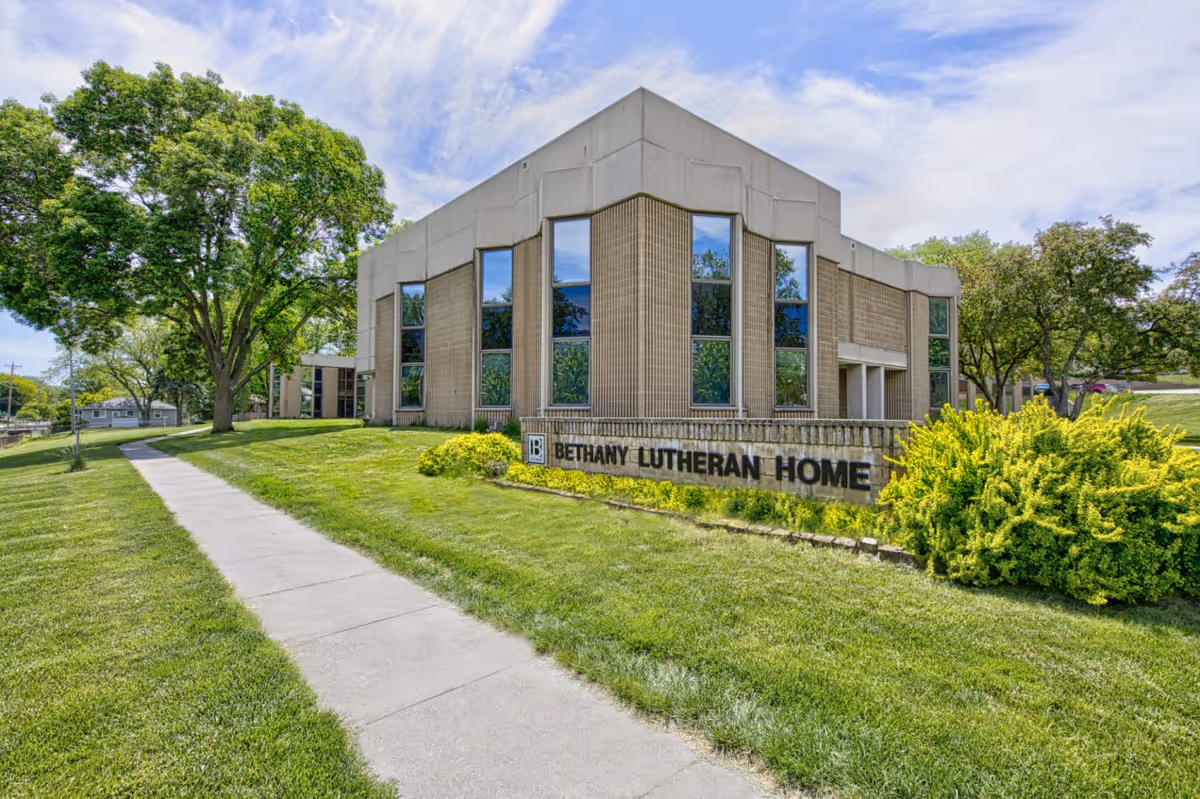 Exterior view of Bethany Lutheran Home building with large windows, surrounded by green grass, trees, and a clear sky. A concrete sidewalk leads up to the building.