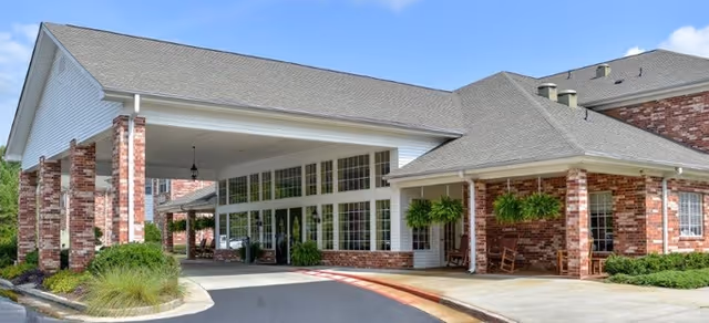 Exterior view of a senior living facility entrance with a covered drop-off area supported by brick columns, large windows, hanging plants, and a clear blue sky.