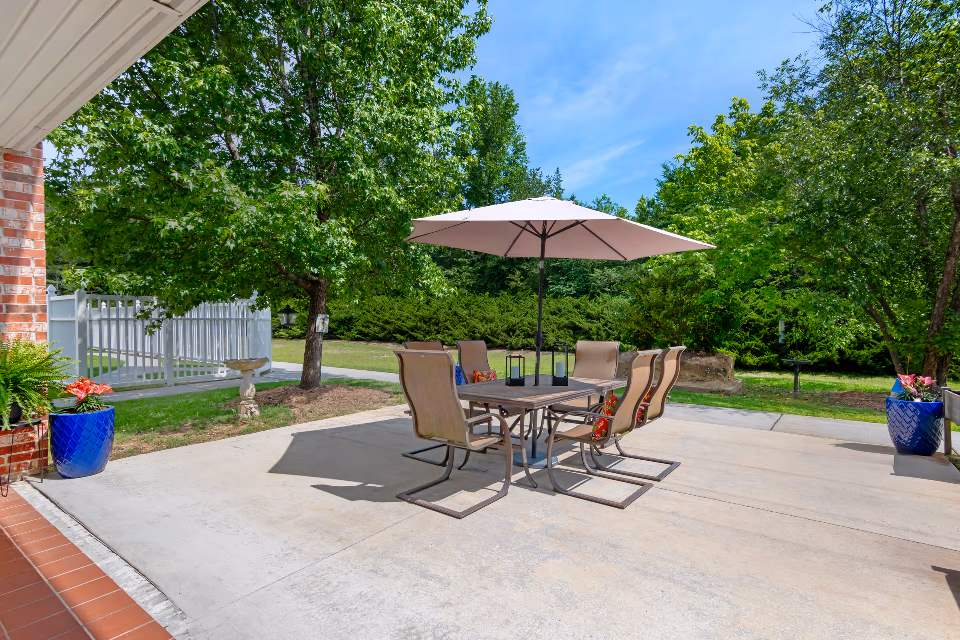 Sunny outdoor patio with a dining table, umbrella and chairs on a concrete slab surrounded by trees and potted plants.