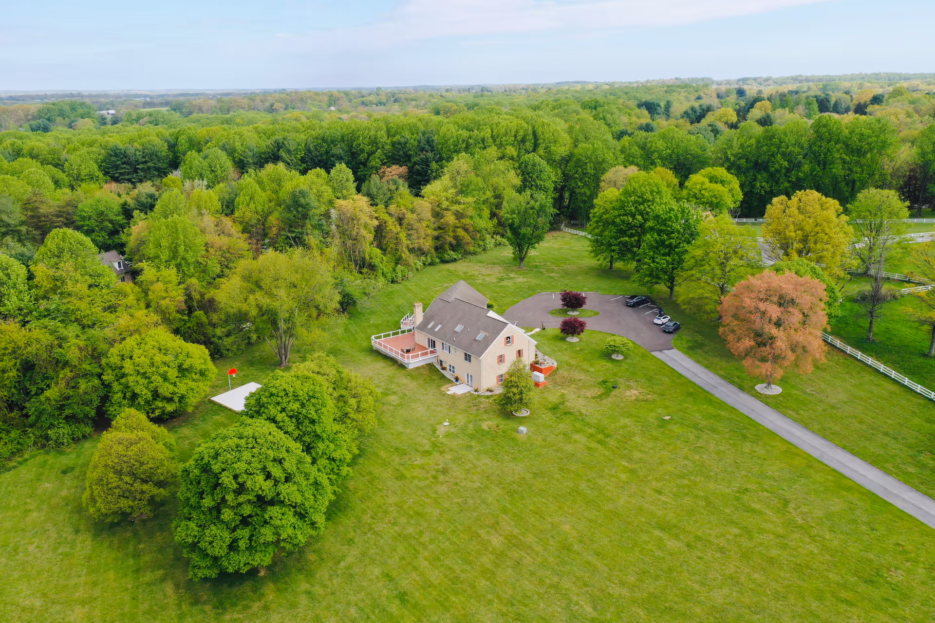 Aerial view of a large house surrounded by expansive green lawns and numerous trees, with a circular driveway and several parked cars. The setting is rural with dense forest in the background under a clear sky.