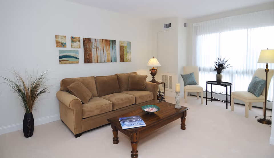Bright furnished living room with a brown sofa, wooden coffee table, chairs by a large window, and wall art.