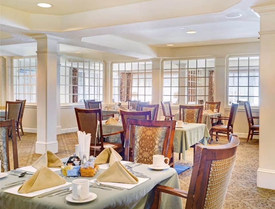 A bright and elegant dining room with multiple tables covered in light green tablecloths, each set with beige folded napkins, white cups and saucers, and silverware. The room features wooden chairs with patterned upholstery, large windows with blinds, and decorative columns and glass panel dividers.