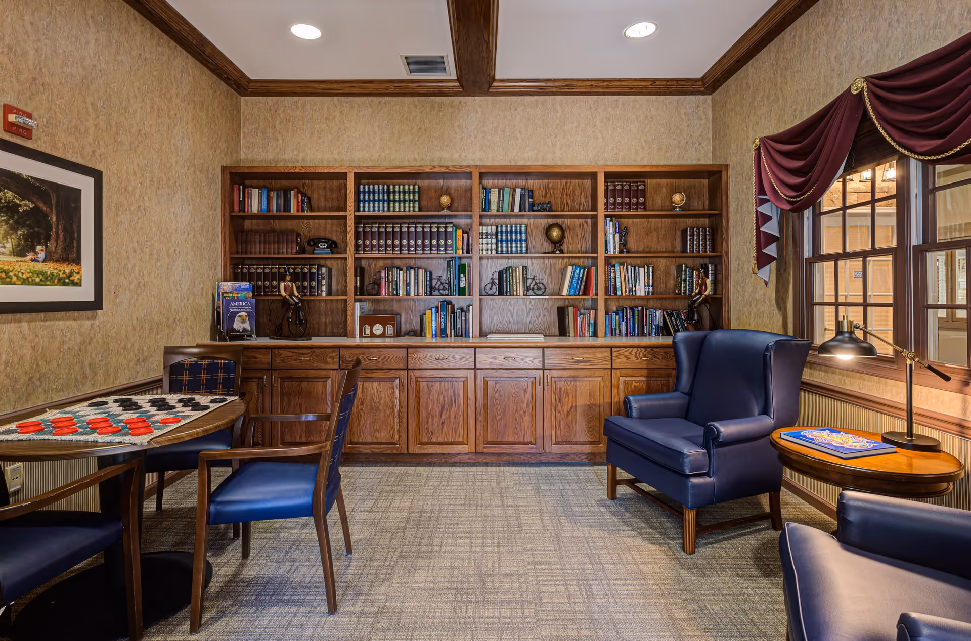 A cozy reading and game room with wooden bookshelves filled with books and decorative items. There are two blue leather armchairs, a round wooden side table with a lamp and a book, and a table with a checkers game set up. The room has beige textured walls, a framed picture on the left wall, and a window with burgundy curtains on the right.