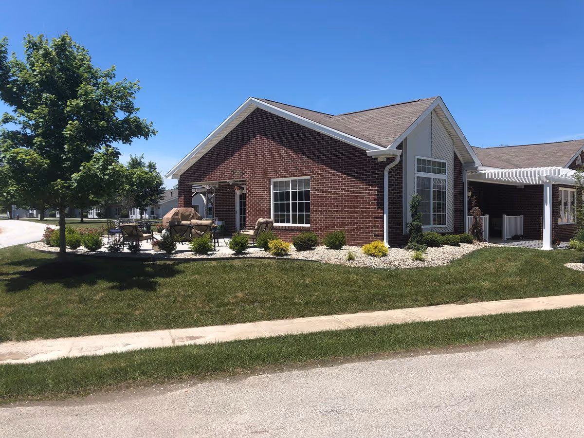 Exterior view of a single-story brick building with a patio area featuring outdoor furniture and a grill. The building is surrounded by a well-maintained lawn, small shrubs, and a tree under a clear blue sky.
