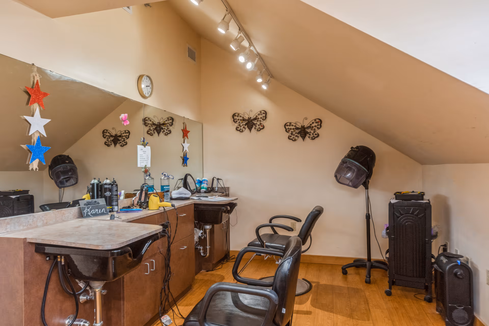 Interior view of a small hair salon area with two black salon chairs, a countertop with hair care products and tools, a large mirror on the wall, decorative butterfly wall hangings, and a hair dryer hood on a stand. The room has slanted ceilings and wooden flooring.