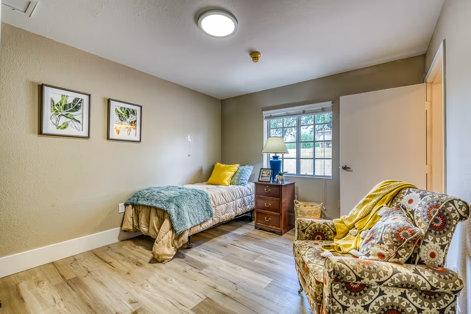 A cozy bedroom with a single bed covered in beige bedding and a teal throw blanket, a wooden nightstand with a lamp and framed photos, a window with a view of greenery outside, and a patterned armchair with a yellow throw blanket and matching pillow. Two framed botanical prints hang on the beige wall.