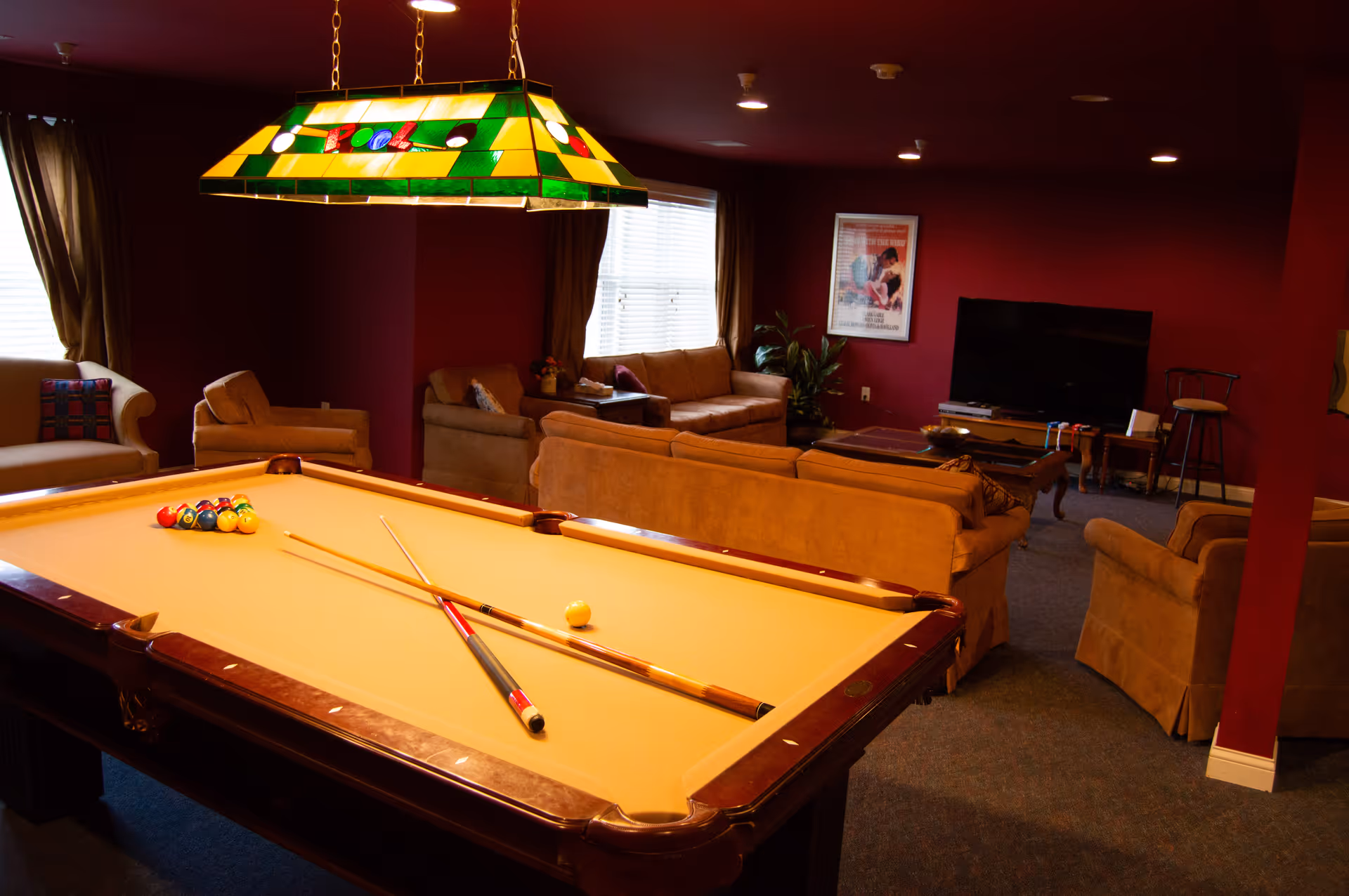 A cozy game and lounge room with a pool table in the foreground, two pool cues resting on the table, and a set of billiard balls arranged for a game. The room has red walls, multiple beige sofas and armchairs, a large flat-screen TV, a coffee table, and a stained glass hanging light fixture above the pool table. Windows with curtains allow natural light into the space.