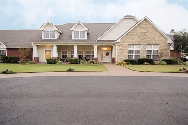 Front exterior view of a single-story residential-style building with a stone facade, multiple windows, a covered porch with chairs, and a well-maintained lawn and sidewalk leading to the entrance.