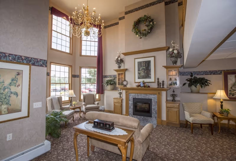 A cozy living room area with high ceilings and large windows letting in natural light. The room features a chandelier, a fireplace with decorative items on the mantel, comfortable armchairs, a loveseat, side tables with lamps, framed artwork on the walls, and plants adding greenery to the space.