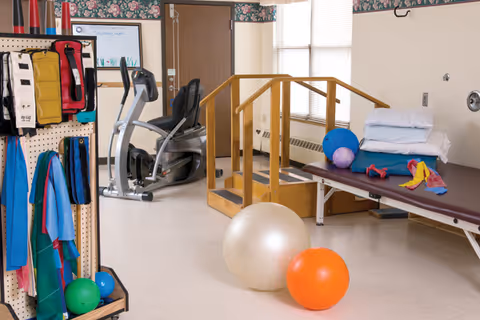 Physical therapy room with exercise equipment including an exercise bike, therapy balls, resistance bands, a wooden handrail ramp, and a padded therapy table with pillows. The room has floral wallpaper border near the ceiling and a window with blinds.