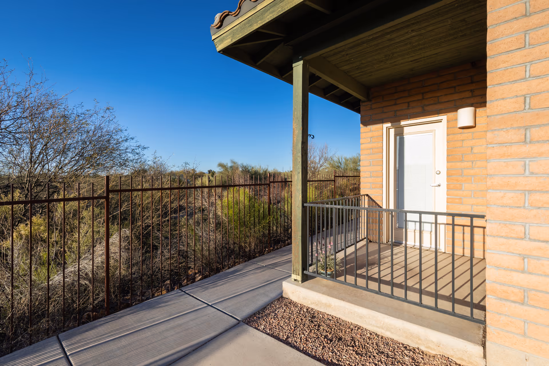 A small outdoor patio area attached to a brick building with a white door and a metal railing. The patio overlooks a natural landscape with dry bushes and trees under a clear blue sky.