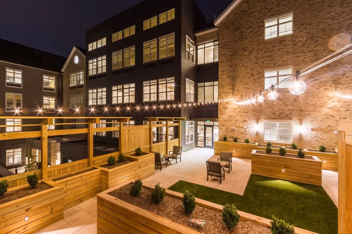 Nighttime view of an outdoor courtyard at Tribute at One Loudoun featuring wooden planter boxes with small shrubs, string lights hanging overhead, several chairs arranged for seating, and surrounding multi-story buildings with illuminated windows.
