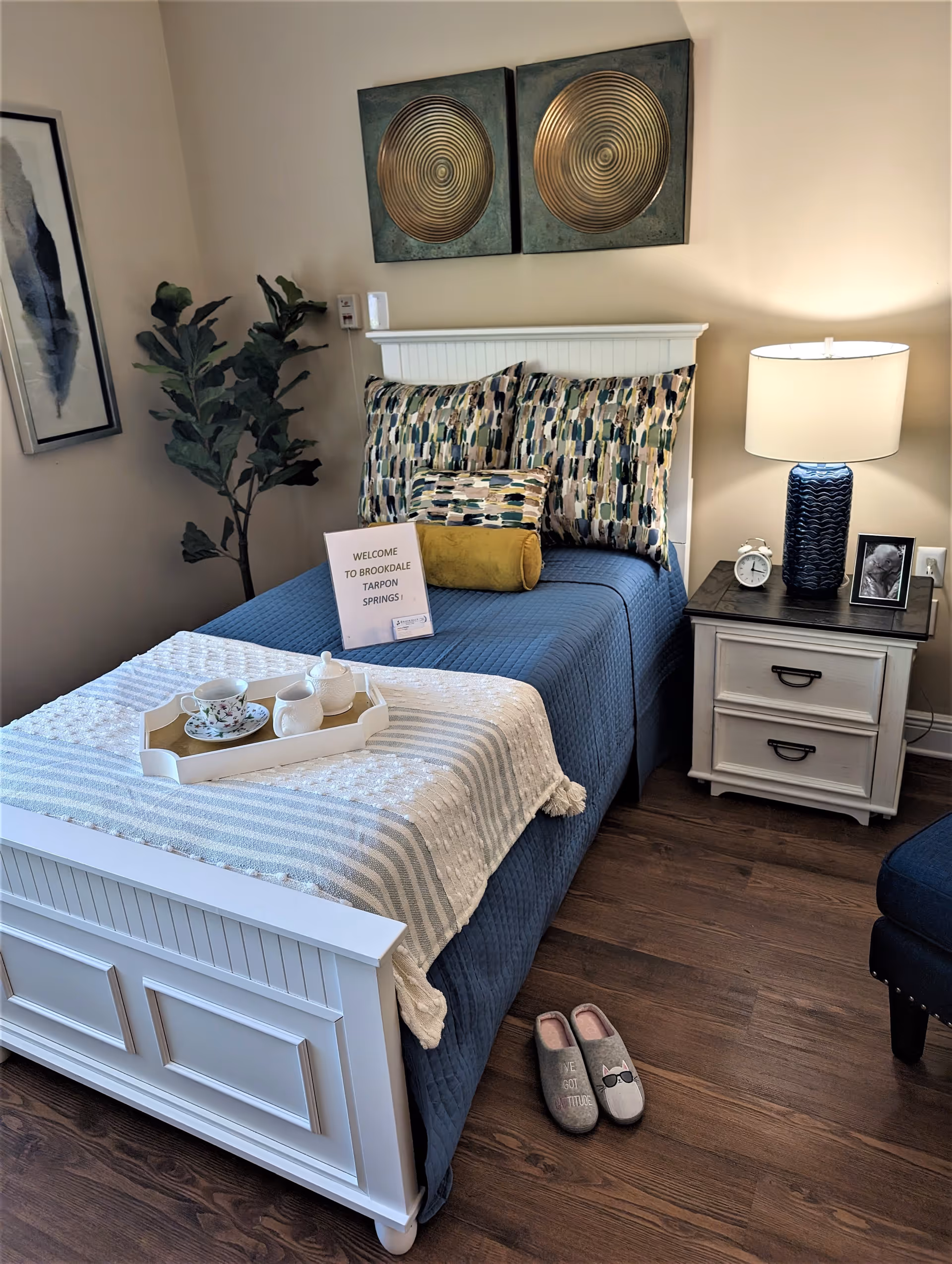 A neatly made single bed with a blue quilt and patterned pillows in a cozy bedroom. A white tray with a teacup and teapot is placed on the bed. Next to the bed is a white nightstand with a blue lamp, a small clock, and a framed photo. A pair of slippers is on the wooden floor beside the bed. There is a plant in the corner and decorative wall art above the bed. A sign on the bed reads 'WELCOME TO BROOKDALE TARPON SPRINGS!'