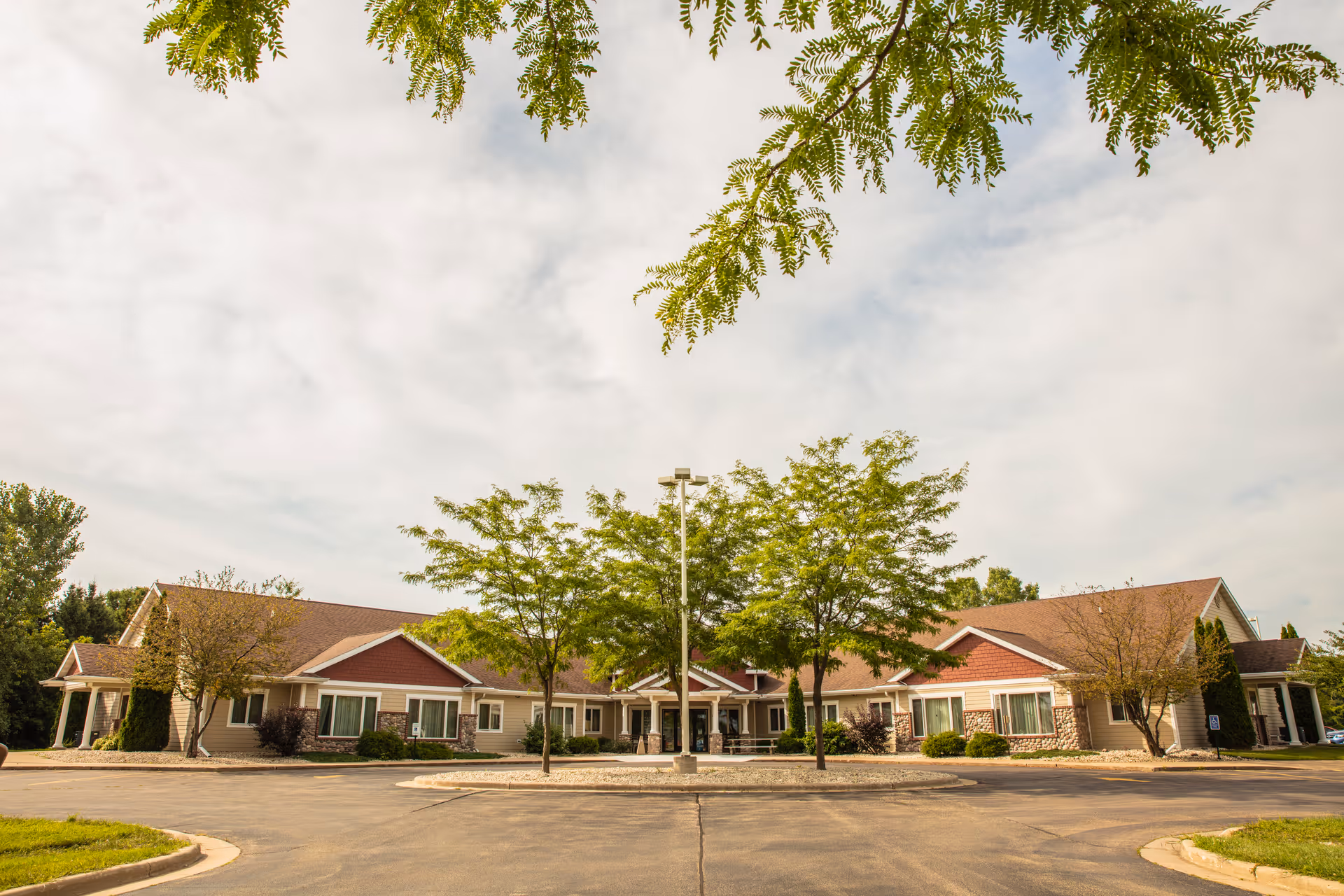 Exterior view of a single-story senior living facility building with a U-shaped layout, surrounded by trees and a paved driveway in front under a partly cloudy sky.
