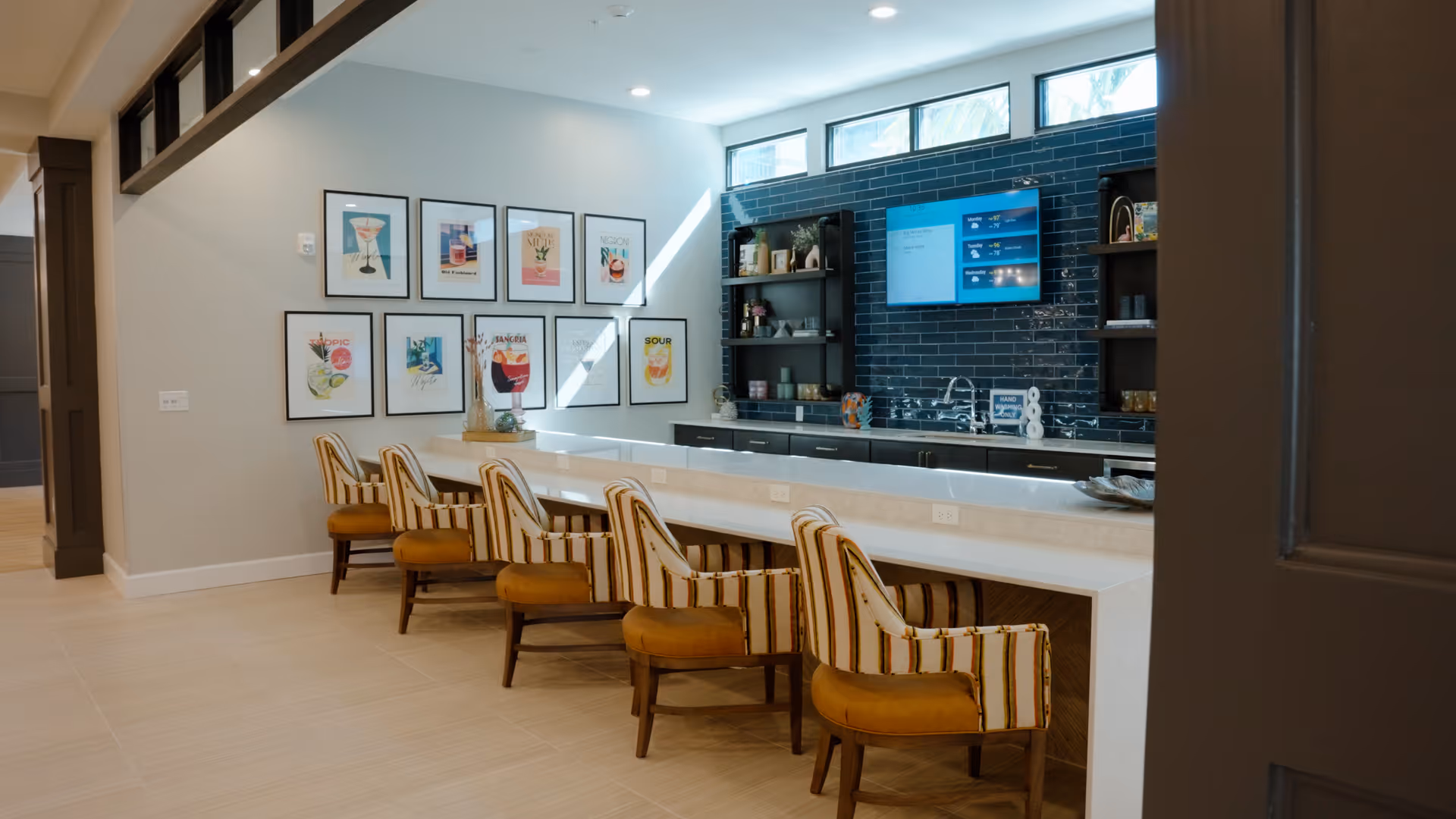 Interior view of a modern kitchen area with a long white countertop and six striped cushioned chairs. The back wall features dark blue subway tiles, open shelving with decorative items, a sink, and a mounted flat-screen TV. The adjacent wall has a series of colorful framed artwork. The space is well-lit with recessed ceiling lights and natural light from high windows.