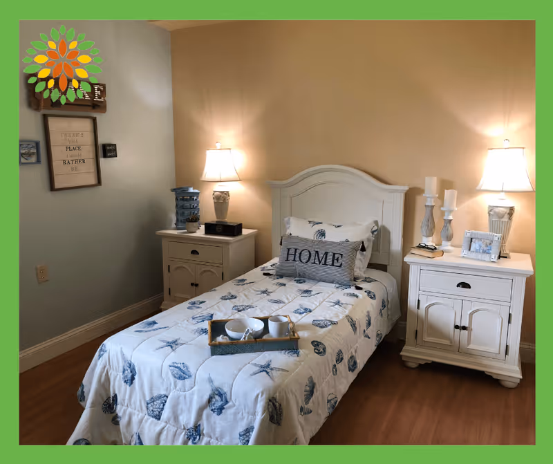 A cozy bedroom with a single bed featuring a white headboard and a seashell-themed quilt. There are two white nightstands on either side of the bed, each with a lamp and decorative items. A pillow with the word 'HOME' is placed on the bed, and a tray with a bowl and cup rests on the quilt. The walls are painted in two tones, beige and light blue, with framed wall art and a small shelf on the left side.