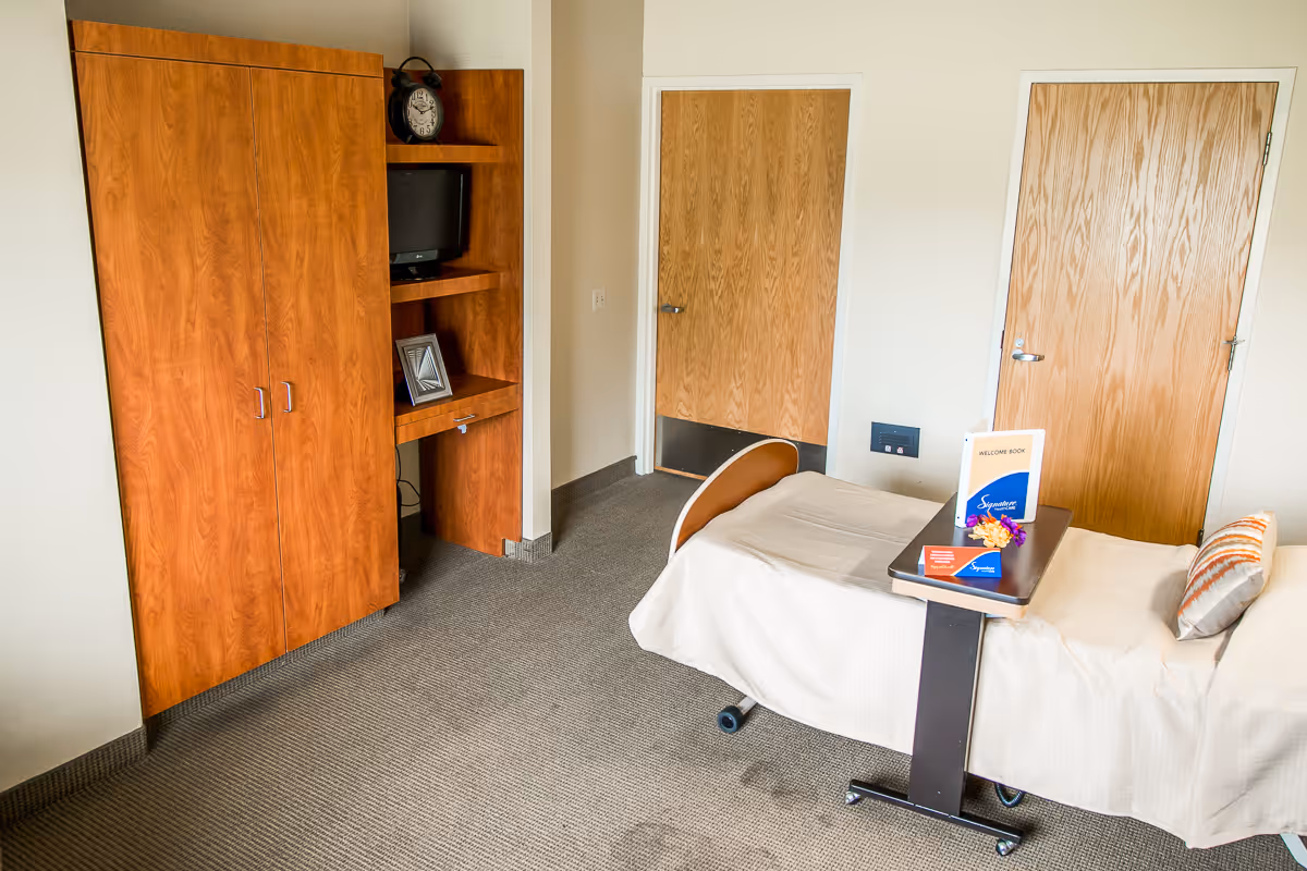 A simple room with a single bed covered with a white sheet and a pillow, a wooden cabinet with shelves holding a small TV, a clock, and a picture frame. There are two wooden doors on the far wall, and a small overbed table with a welcome book and flowers on it.