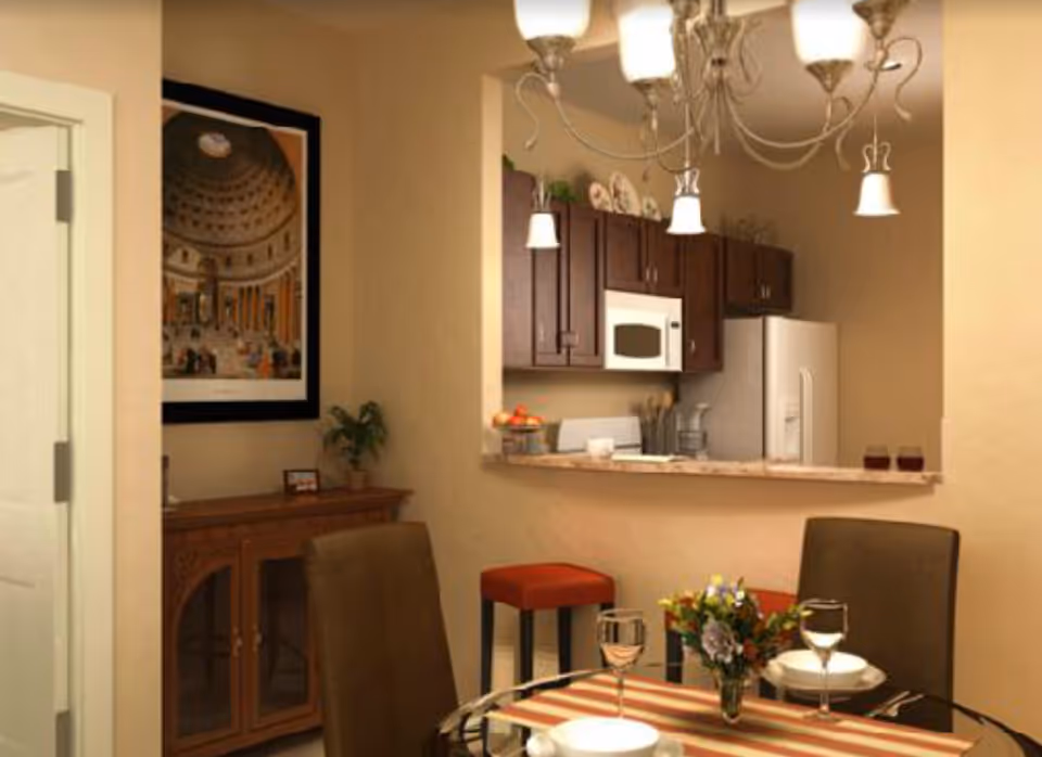 Interior view of a dining area and kitchen in a senior living facility. The dining area has a round glass table set with plates, wine glasses, and a small flower arrangement. There are two brown chairs and a red stool near the kitchen counter. The kitchen features dark wooden cabinets, a white microwave, and a white refrigerator. A chandelier with multiple lights hangs above the dining table. A framed picture and a small plant are on a wooden sideboard against the wall.
