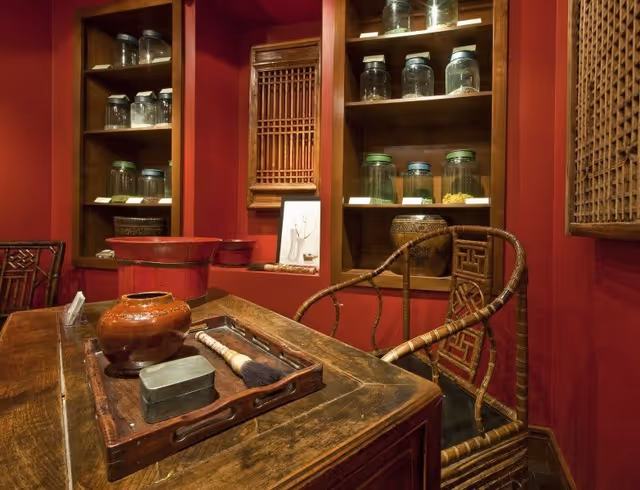 A cozy interior room with red walls featuring wooden shelves holding glass jars and decorative items. A wooden table with a tray containing a brush, a small metal box, and a ceramic pot is in the foreground. A bamboo chair with a woven backrest is positioned next to the table.