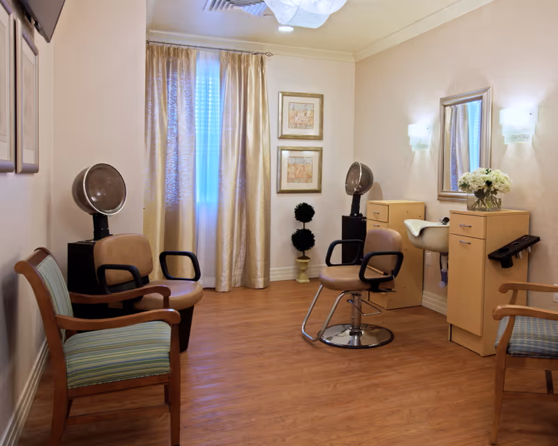 A cozy hair salon room with wooden flooring, two hair drying chairs with hooded dryers, a salon chair in front of a mirror and sink, two wooden armchairs with striped cushions, beige curtains covering a window, two framed pictures on the wall, and a small decorative plant in the corner.