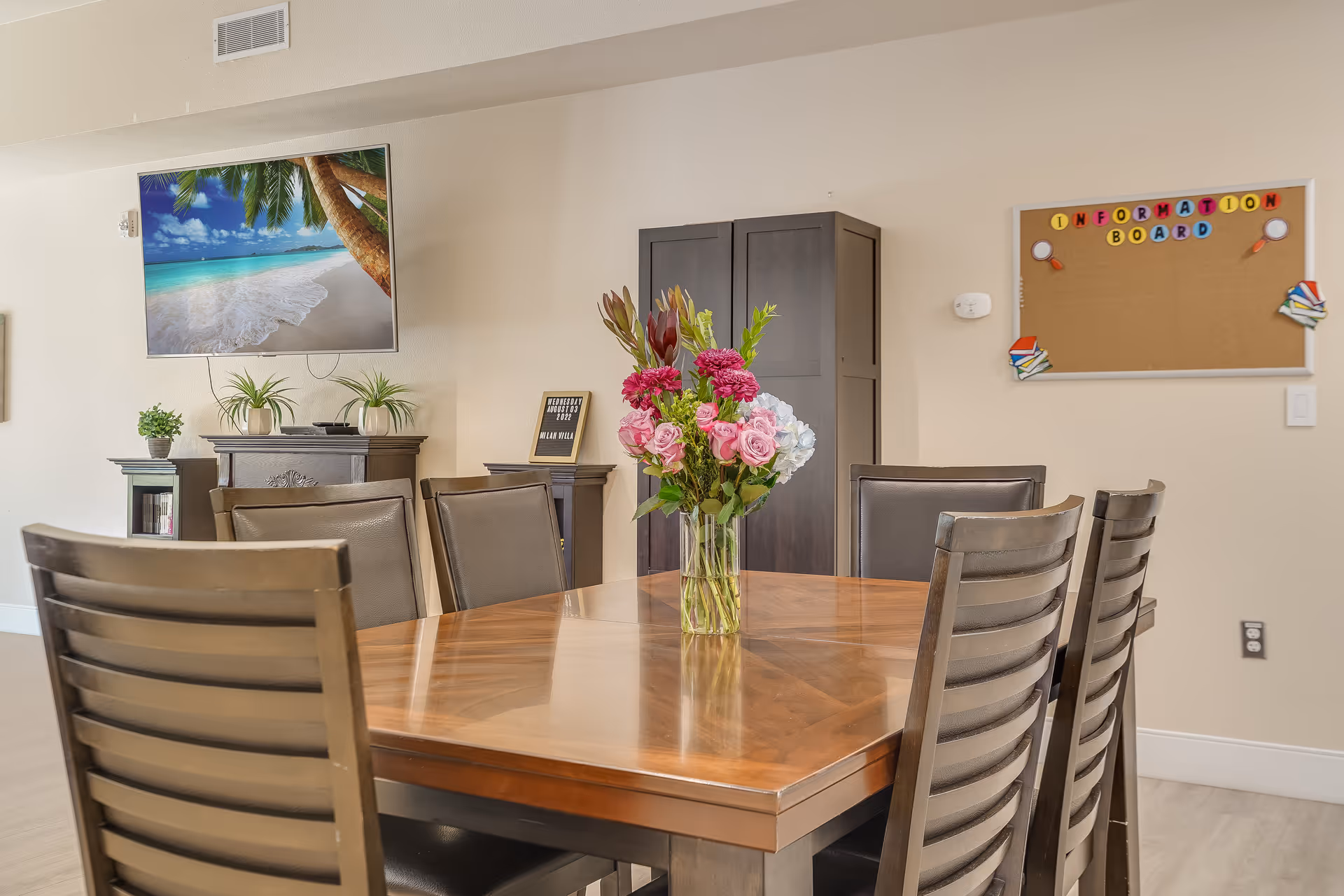 A dining room with a wooden table surrounded by eight chairs. A vase with a colorful bouquet of flowers is placed in the center of the table. On the wall, there is a mounted TV displaying a beach scene, a cork information board with colorful letters, and a small sign on a stand that reads the date and facility name. There are also some small plants on furniture against the wall.