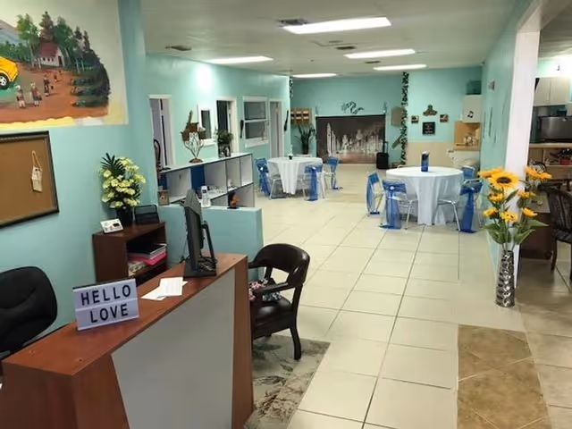 Interior view of a bright and spacious adult daycare facility with light blue walls, tiled floors, and several round tables covered with white tablecloths and surrounded by blue chairs. A reception desk with a sign that reads 'HELLO LOVE' is visible in the foreground, along with decorative plants and wall art.