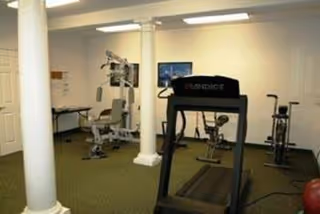 A small fitness room with exercise equipment including a treadmill, stationary bikes, and a multi-function weight machine. The room has white walls, a green carpet, two white columns, and fluorescent ceiling lights.