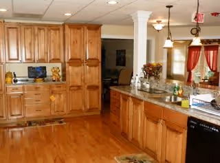 A spacious kitchen with wooden cabinets and drawers, a granite countertop, and hardwood flooring. The kitchen features a dishwasher, a microwave, and a sink area with decorative items and flowers. There are pendant lights hanging from the ceiling and red curtains on the windows in the background.