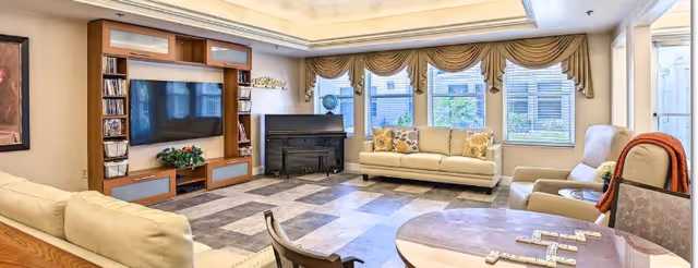 A bright and spacious living room with beige sofas, a wooden entertainment center with a large flat-screen TV, a piano in the corner, and large windows with decorative valances allowing natural light to fill the room. A round table with chairs and a game of dominoes is in the foreground.
