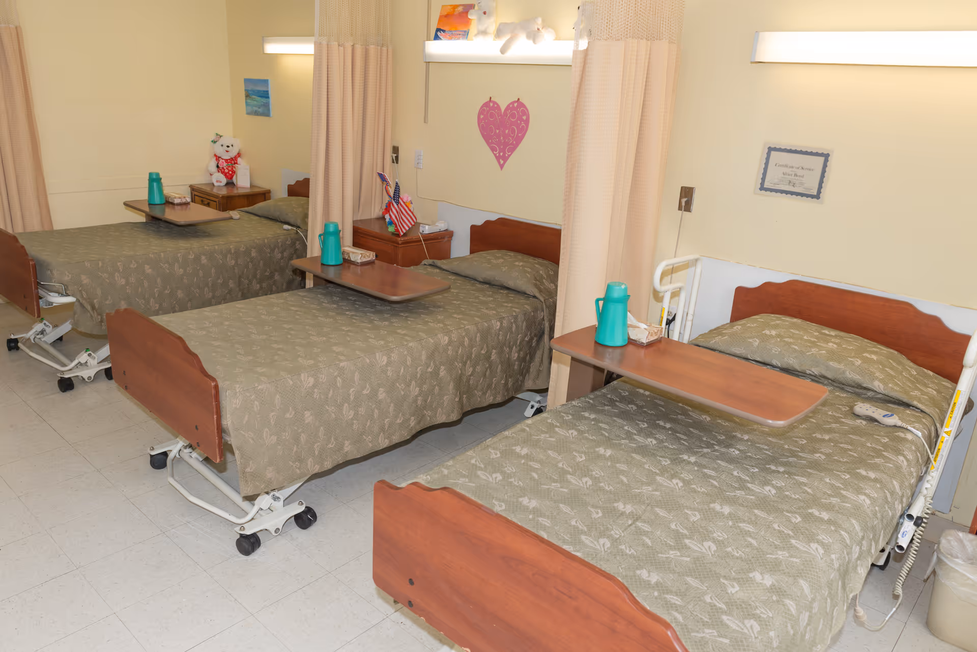 A room with three hospital-style beds arranged side by side, each with a green patterned bedspread and a wooden overbed table. The beds are separated by beige privacy curtains. Each bed has a green water pitcher and a tissue box on the table. The room has light yellow walls with a pink heart decoration and a certificate on the wall. There is a teddy bear on a small table near the farthest bed.