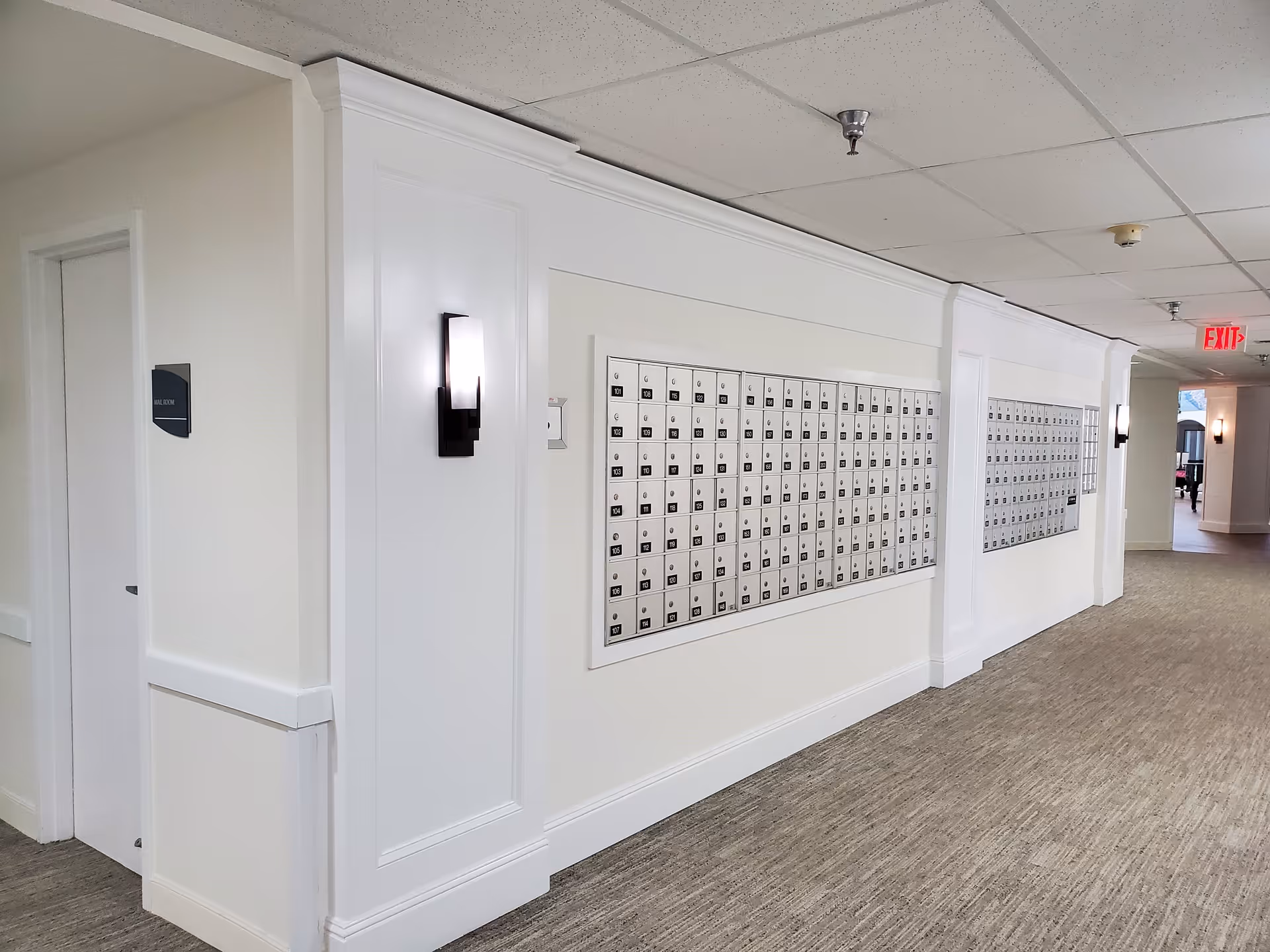 Interior hallway of a senior living facility with a wall of mailboxes and a door labeled 'Mail Room'. The hallway has beige walls, carpeted floor, ceiling lights, and an exit sign in the distance.