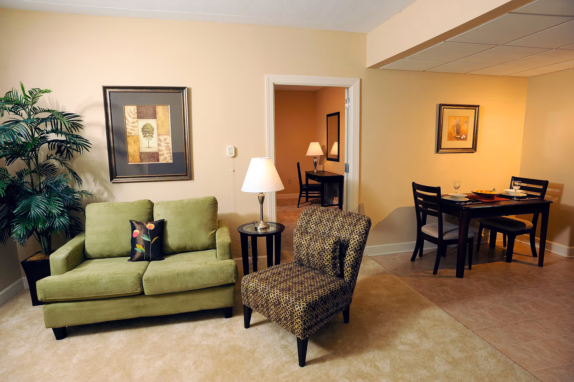 A cozy living area featuring a green loveseat with a decorative pillow, a patterned armchair, a small round side table with a lamp, and a large potted plant. In the background, there is a dining table set for two with plates, glasses, and a bowl, along with framed artwork on the walls. An open doorway reveals another room with a desk, chair, lamp, and mirror.