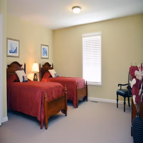 A bedroom with two wooden twin beds covered in red bedspreads, each with a pillow and a decorative cushion. A lamp sits on a small table between the beds. The room has light yellow walls, a window with white blinds, a black chair, and a vase with red and white flowers.