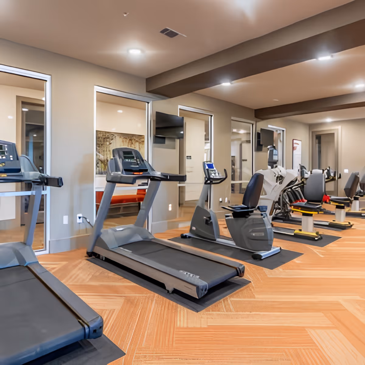 Interior view of a fitness room with treadmills, stationary bikes, and other exercise equipment arranged on a wooden floor with large mirrors on the walls reflecting the equipment.
