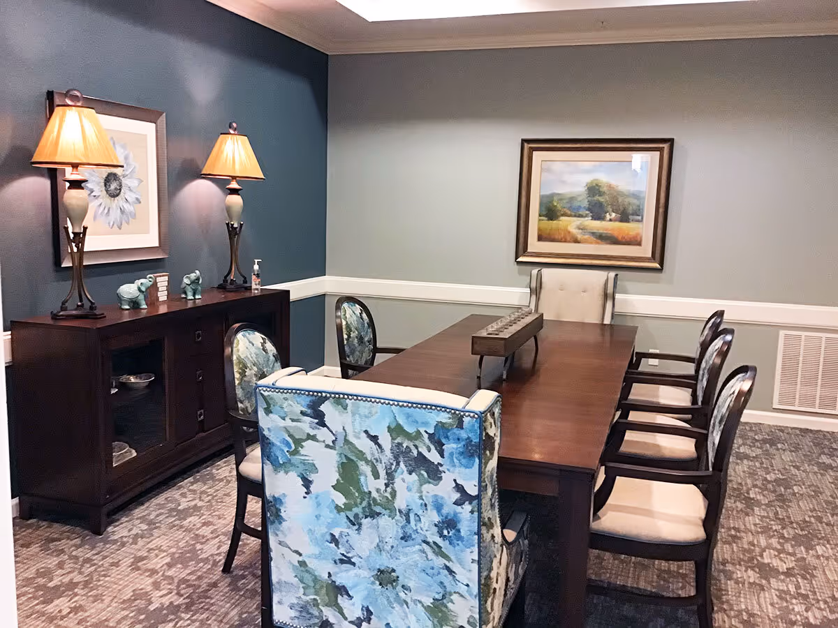 A dining room with a long wooden table surrounded by eight chairs, two of which have floral patterned upholstery. On the left side, there is a dark wooden sideboard with two table lamps, decorative elephant figurines, and a framed flower artwork above it. On the far wall, there is a framed landscape painting. The room has a carpeted floor and walls painted in two tones of blue and gray.