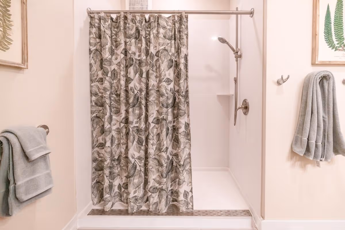 A bathroom shower area with a floral-patterned shower curtain partially drawn to the side, revealing a white shower stall with a handheld showerhead mounted on the wall. Light gray towels hang on hooks and a towel bar on the adjacent walls, which are decorated with framed botanical prints.